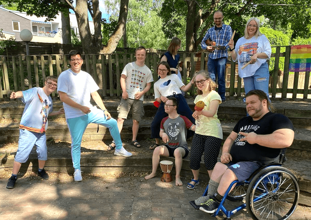Eine Gruppe Jugendlicher mit Down-Syndrom, die fröhlich für das Bild posieren. Mit dabei sind 3 Erzieher und eine Erzieherin. Einer der Erzieher sitzt im Rollstuhl.
